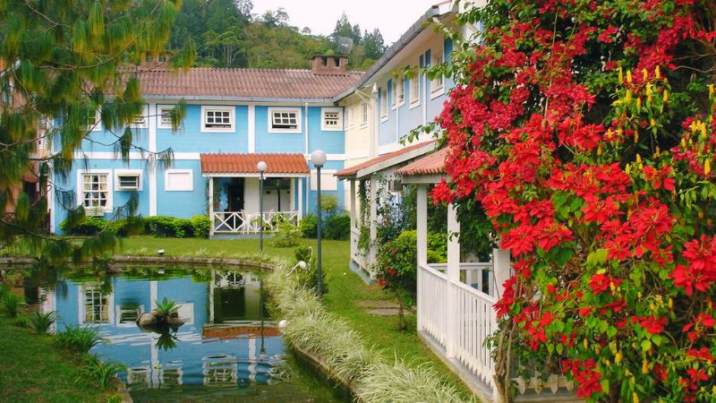 Vista de uma casa azul em frente ao um lago arborizado com flores vermelhas à direita na Pequena Finlândia em Penedo, RJ.